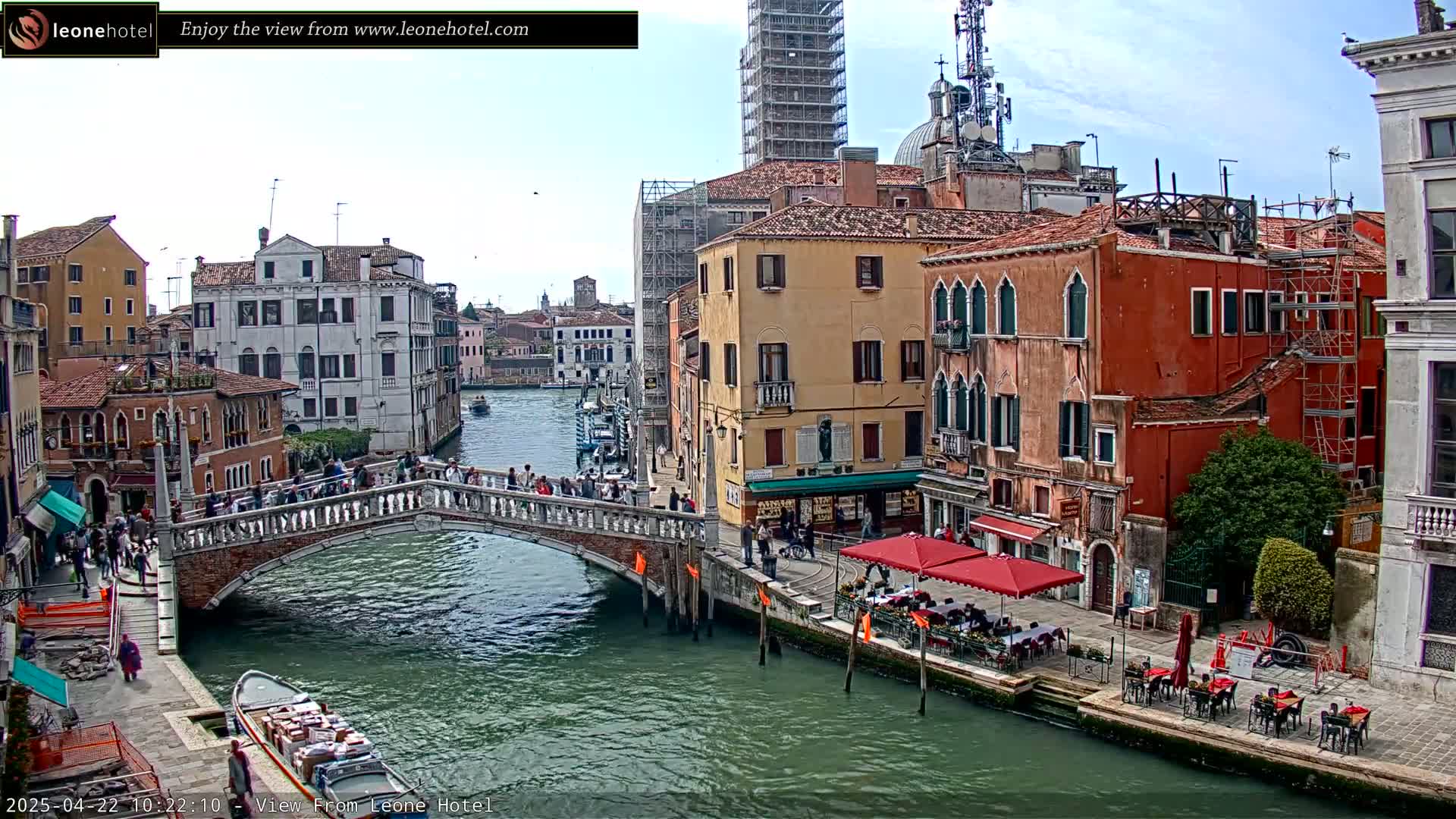 A canal in Venice, Italy, on a partly cloudy day, shows a stone bridge with pedestrians crossing it, colorful buildings lining the waterway, and outdoor seating at a cafe.