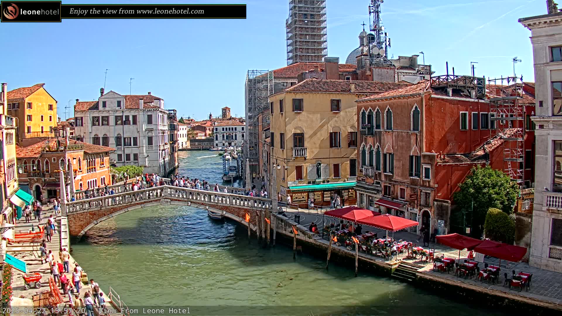 A sunny day in Venice shows a canal with a bridge crowded with people, lined with colorful buildings, some undergoing construction, and outdoor cafes.