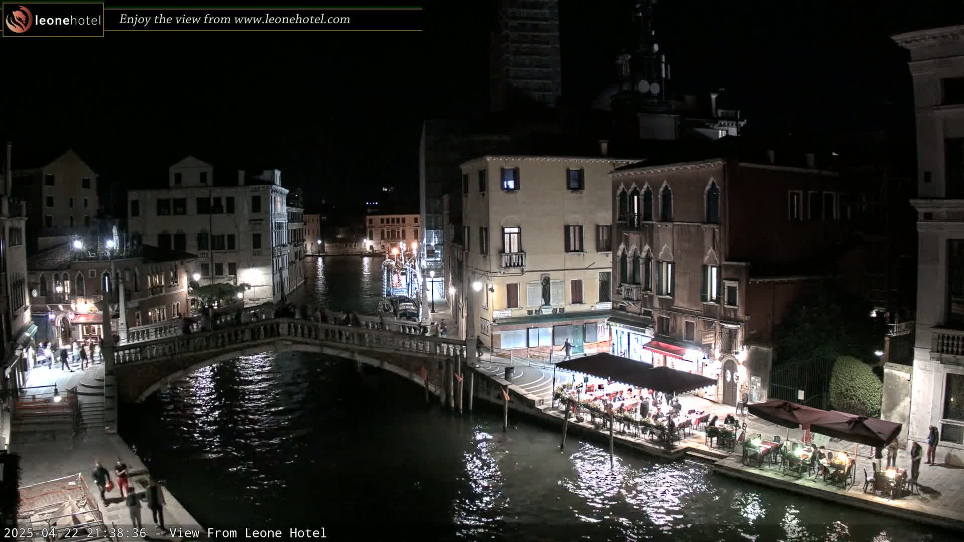 A nighttime view of a Venetian canal, with bridges, buildings, and outdoor restaurants lit along the waterway under a clear sky.