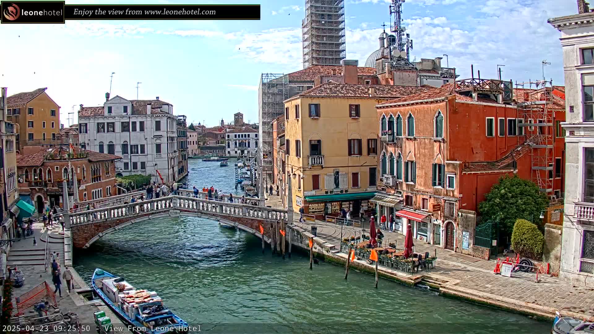 A sunny day in Venice shows a canal with a stone bridge, colorful buildings lining the waterway, and boats moored along the banks.