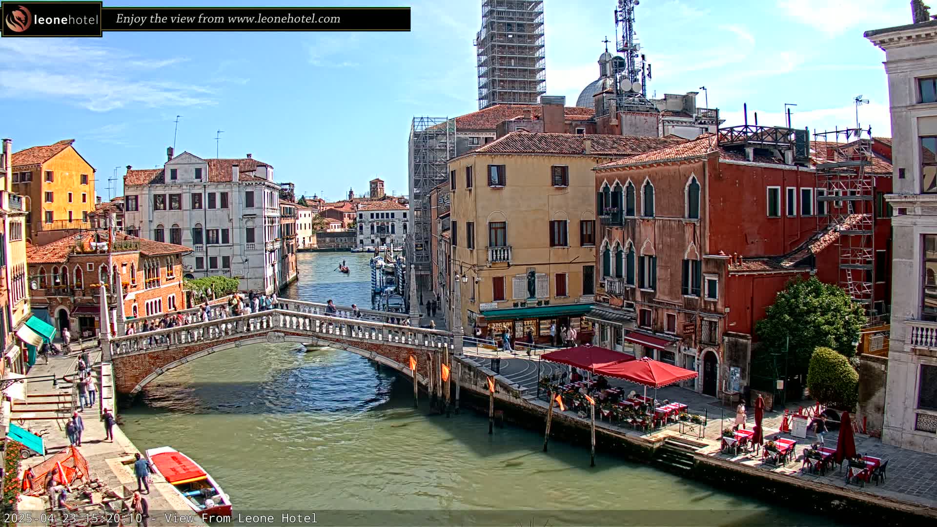 A sunny day in Venice shows a canal with a bridge, lined with colorful buildings, some with scaffolding, and outdoor seating at cafes.