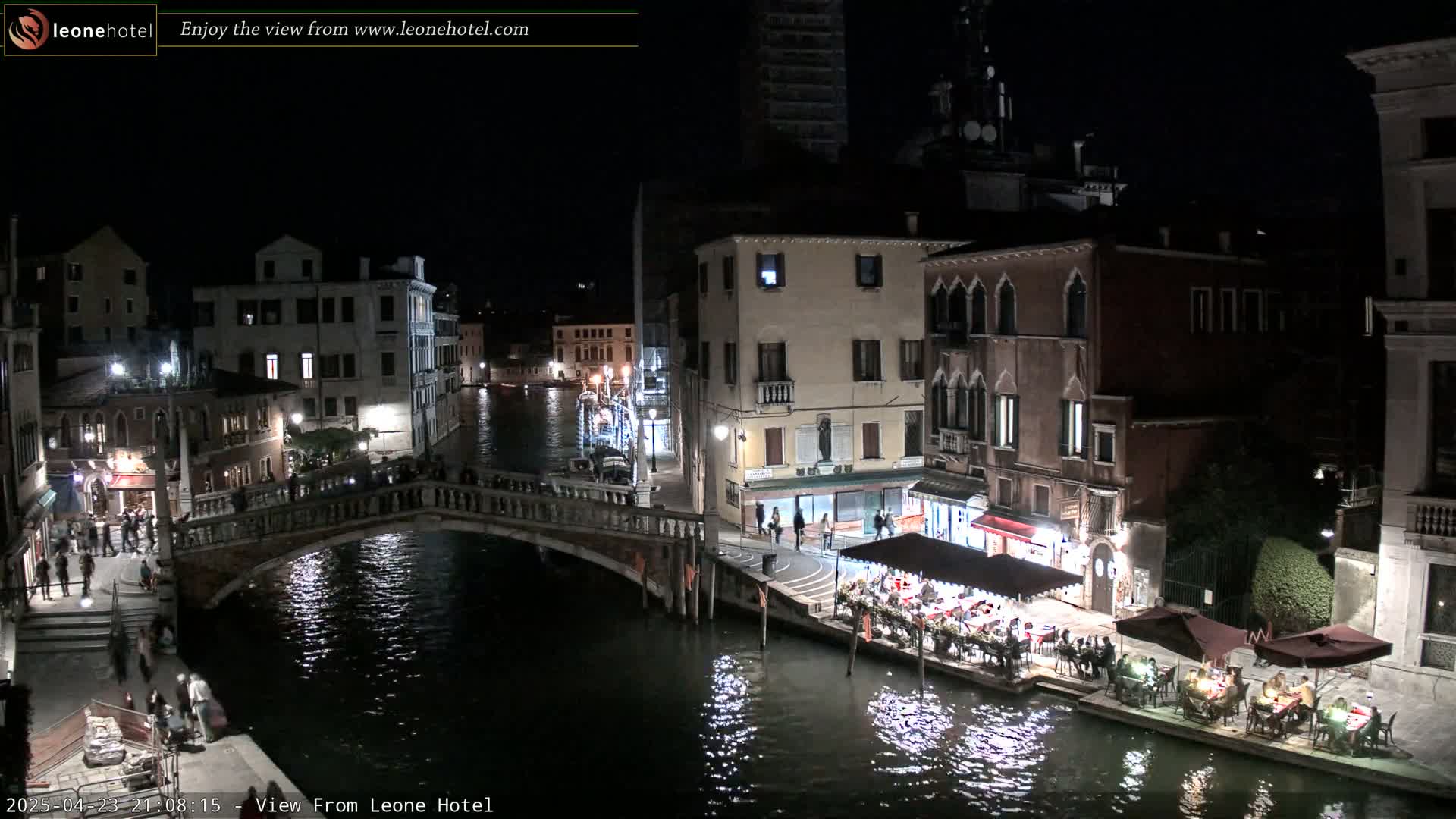 A nighttime view of a canal in Venice, Italy, shows several buildings, a bridge with people on it, and outdoor dining areas along the water's edge.