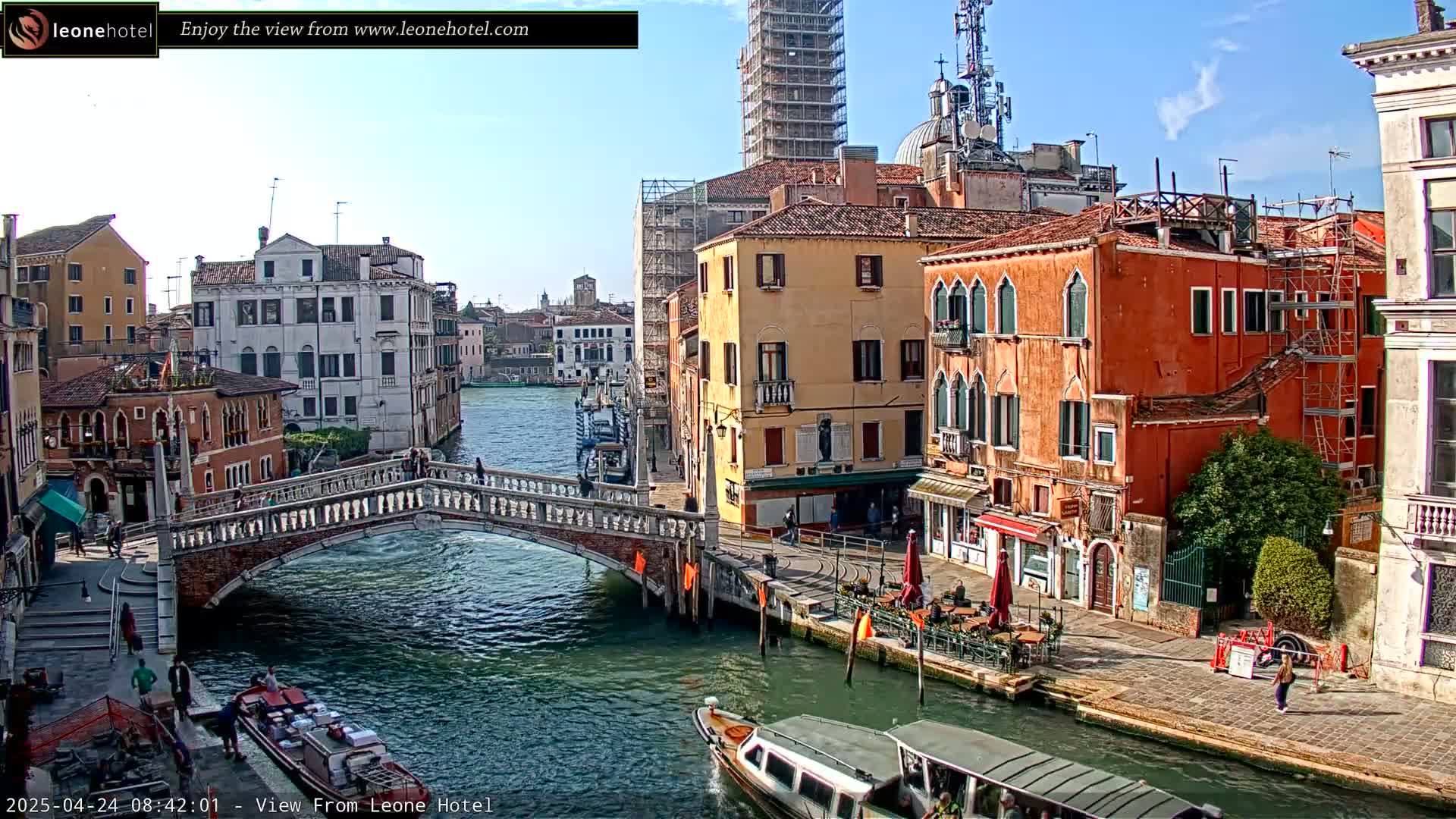 A sunny day in Venice shows a canal with a stone bridge, colorful buildings, boats, and pedestrians.
