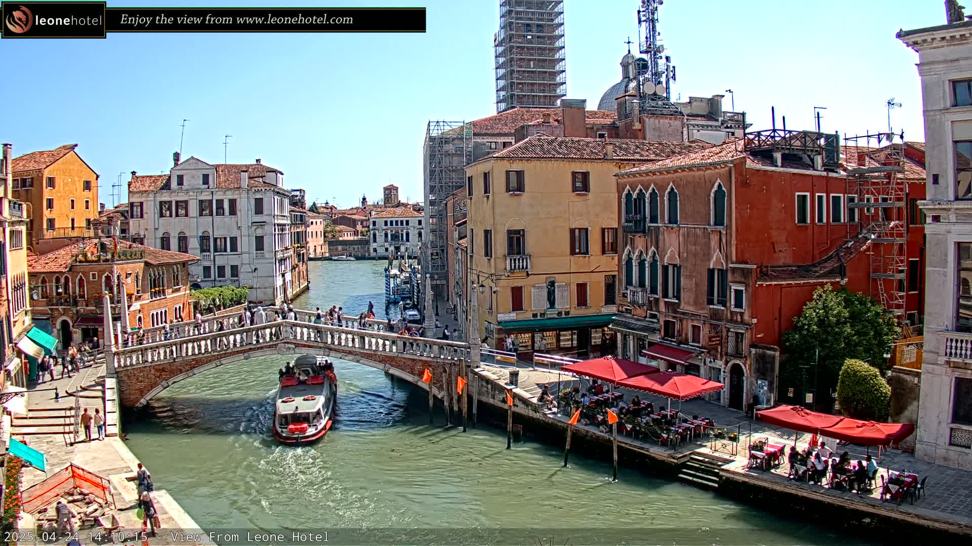 A sunny day in Venice shows a canal with a stone bridge, boats, and people dining outdoors at cafes along the waterway, with multi-story buildings lining the canal.