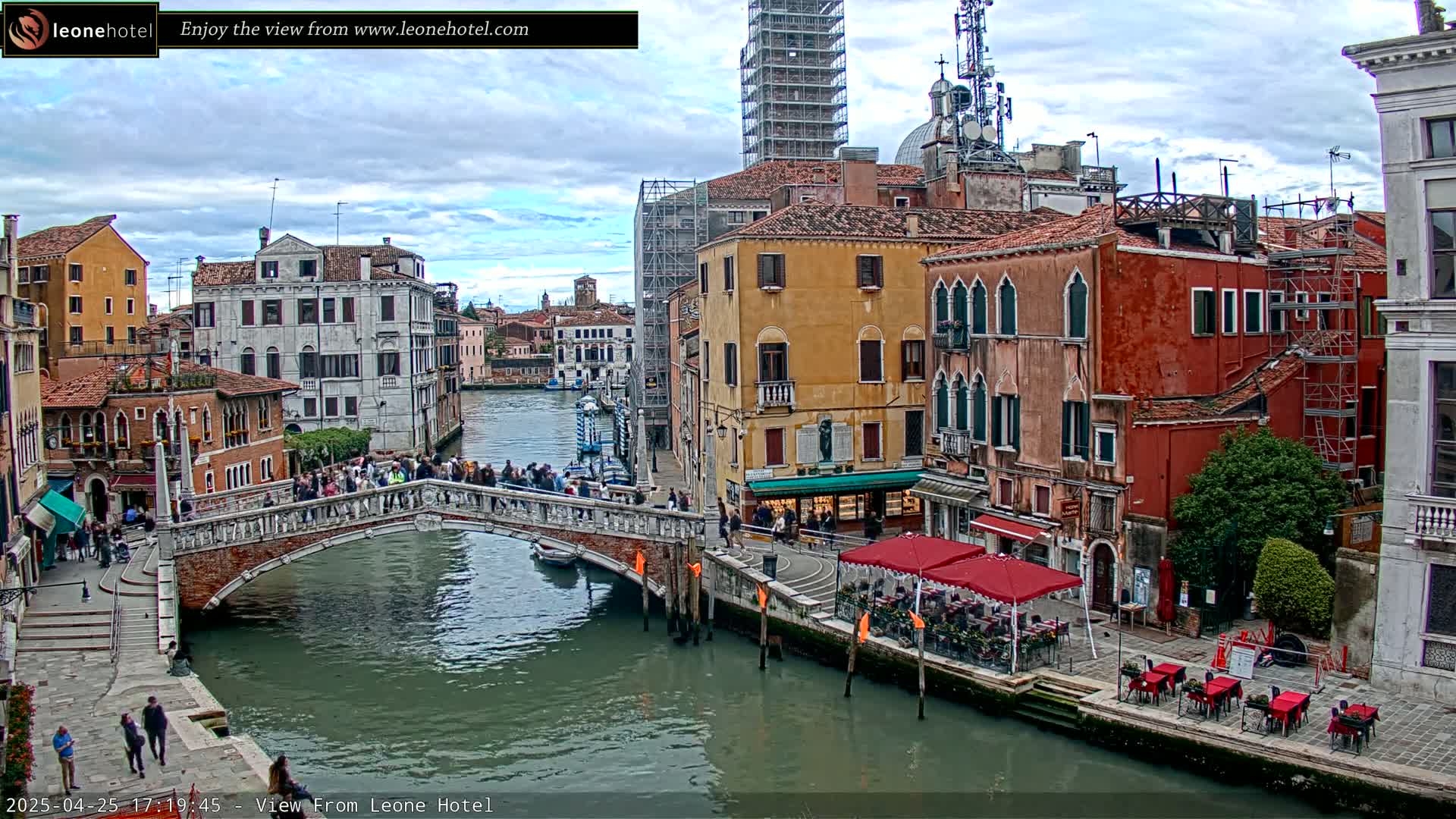 A canal in Venice, under a partly cloudy sky, is spanned by a bridge crowded with people, with colorful buildings lining the waterway and outdoor seating at cafes along the shore.