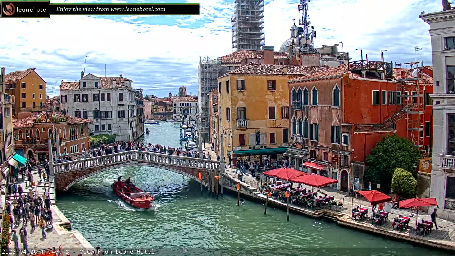 A canal in Venice, under a partly cloudy sky, shows a stone bridge crowded with people, boats on the water, and outdoor restaurants along the canal-side.