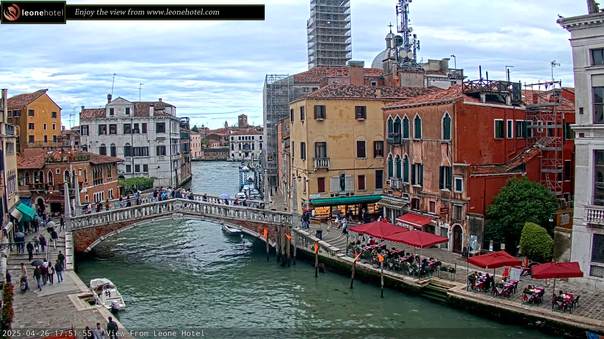 A canal in Venice, Italy, with a stone bridge, colorful buildings, and outdoor seating under red umbrellas, on a cloudy day.