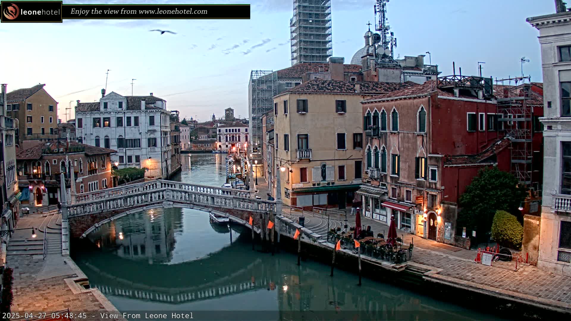 A tranquil canal scene in Venice at dawn shows several buildings lining the waterway, a stone bridge arching over the water, and a few boats moored along the banks.