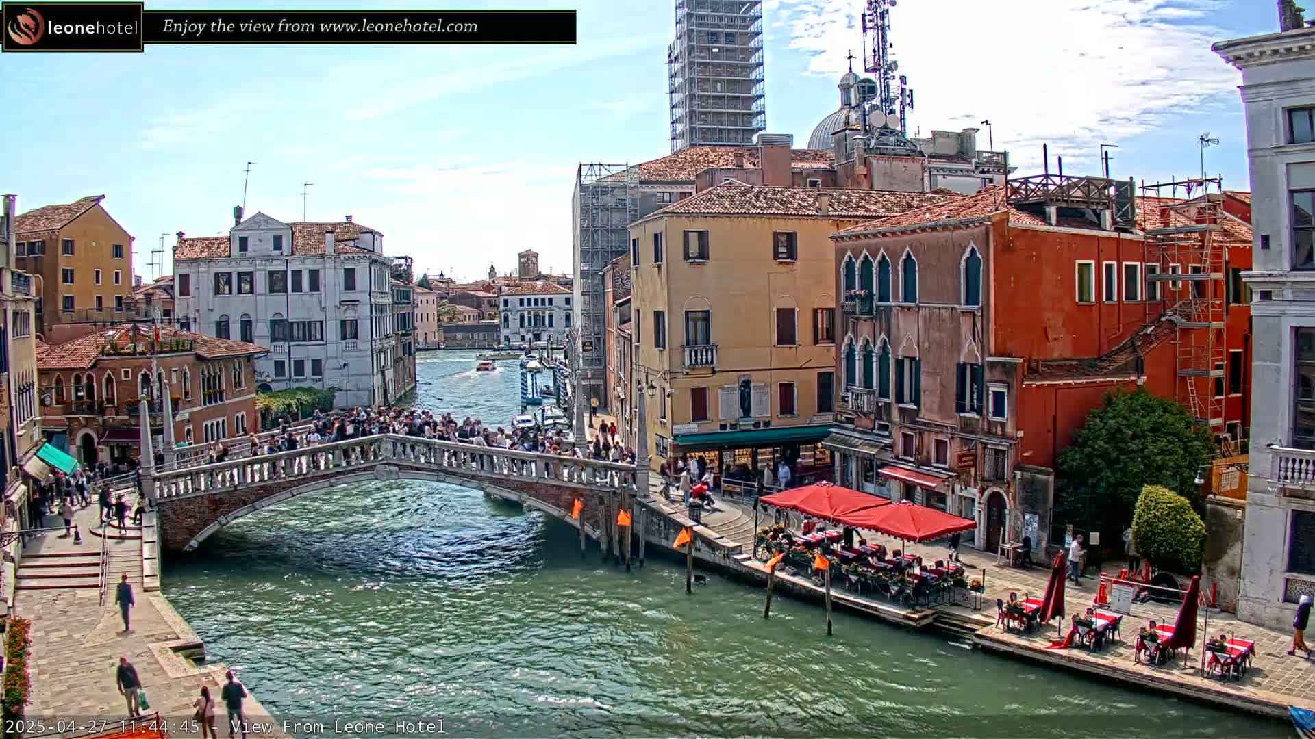 A sunny day in Venice shows a canal with a bridge crowded with people, colorful buildings lining the waterway, and outdoor seating at a restaurant.