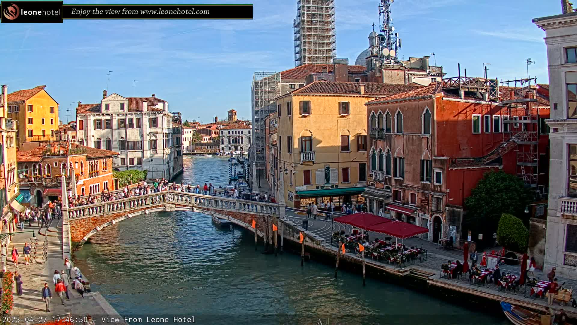 A sunny Venetian canal scene shows a stone bridge crowded with people, colorful buildings lining the waterway, and outdoor cafes with patrons seated at tables.