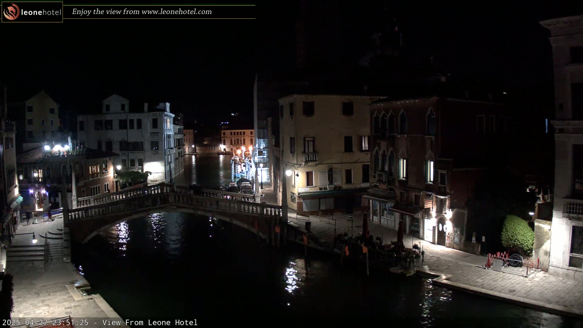 A nighttime view of a canal in Venice, Italy, shows a stone bridge over dark water, with buildings lining the canal on either side, lit by streetlights.