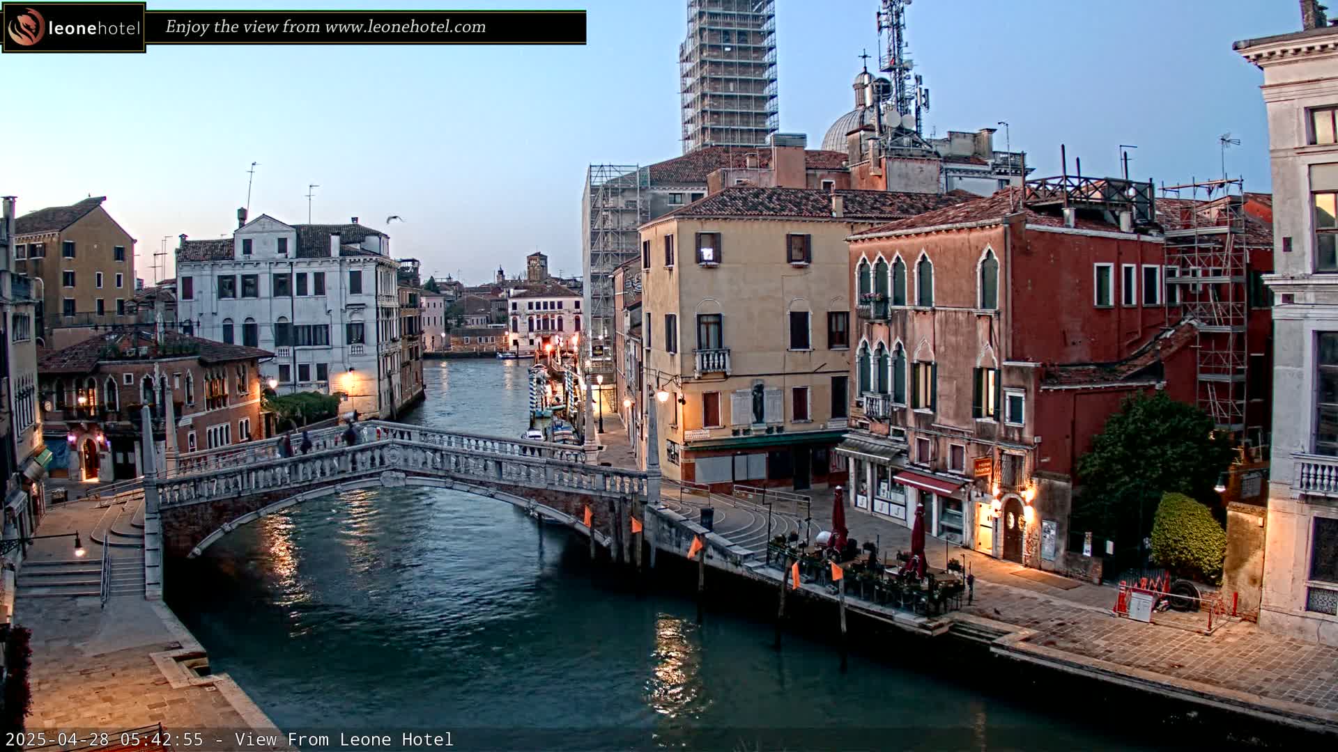 A stone bridge crosses a calm canal in Venice at twilight, with colorful buildings lining the waterway and a few people walking across the bridge.