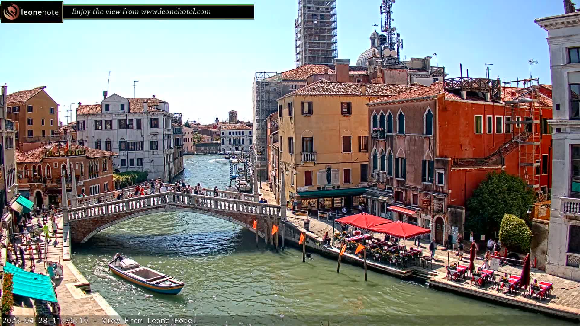 A sunny day in Venice shows a canal with a stone bridge, colorful buildings lining the waterway, and people walking on the bridge and sitting at outdoor cafes.