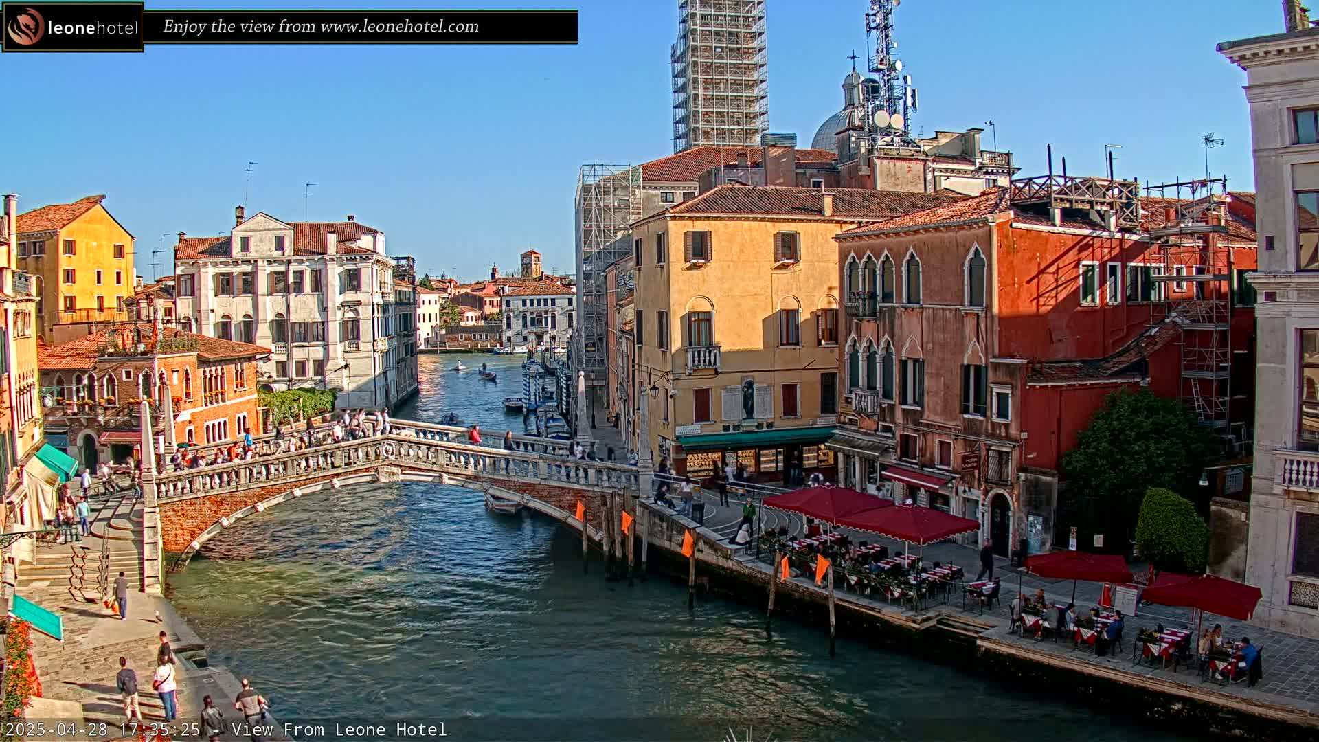 A sunny day in Venice shows a canal with gondolas and a stone bridge, lined with colorful buildings, some with outdoor seating.
