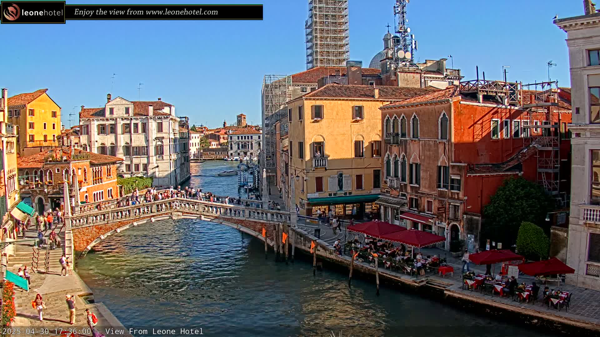 A sunny day in Venice shows a canal with a bridge crowded with people, colorful buildings lining the waterway, and outdoor seating at a restaurant.