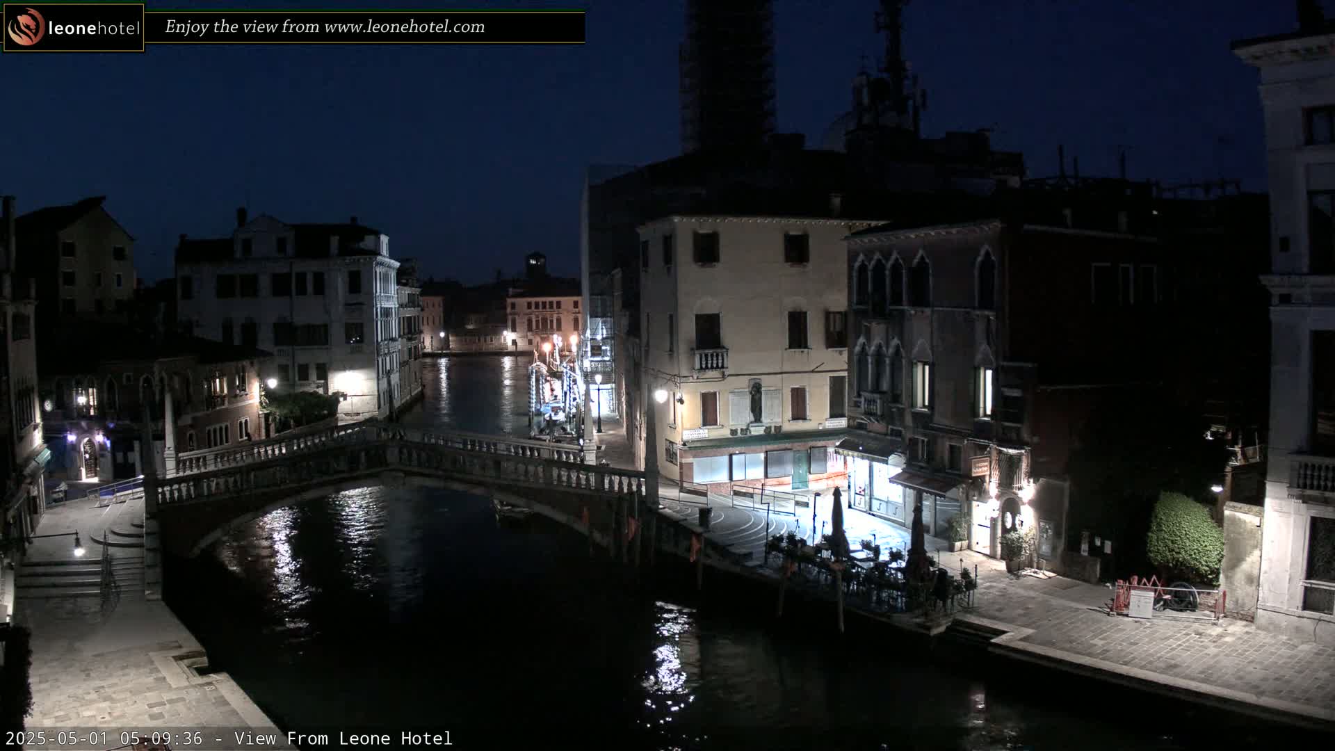 A nighttime view of a canal in Venice, Italy, shows a bridge, buildings lining the waterway, and a few people sitting outside a restaurant.