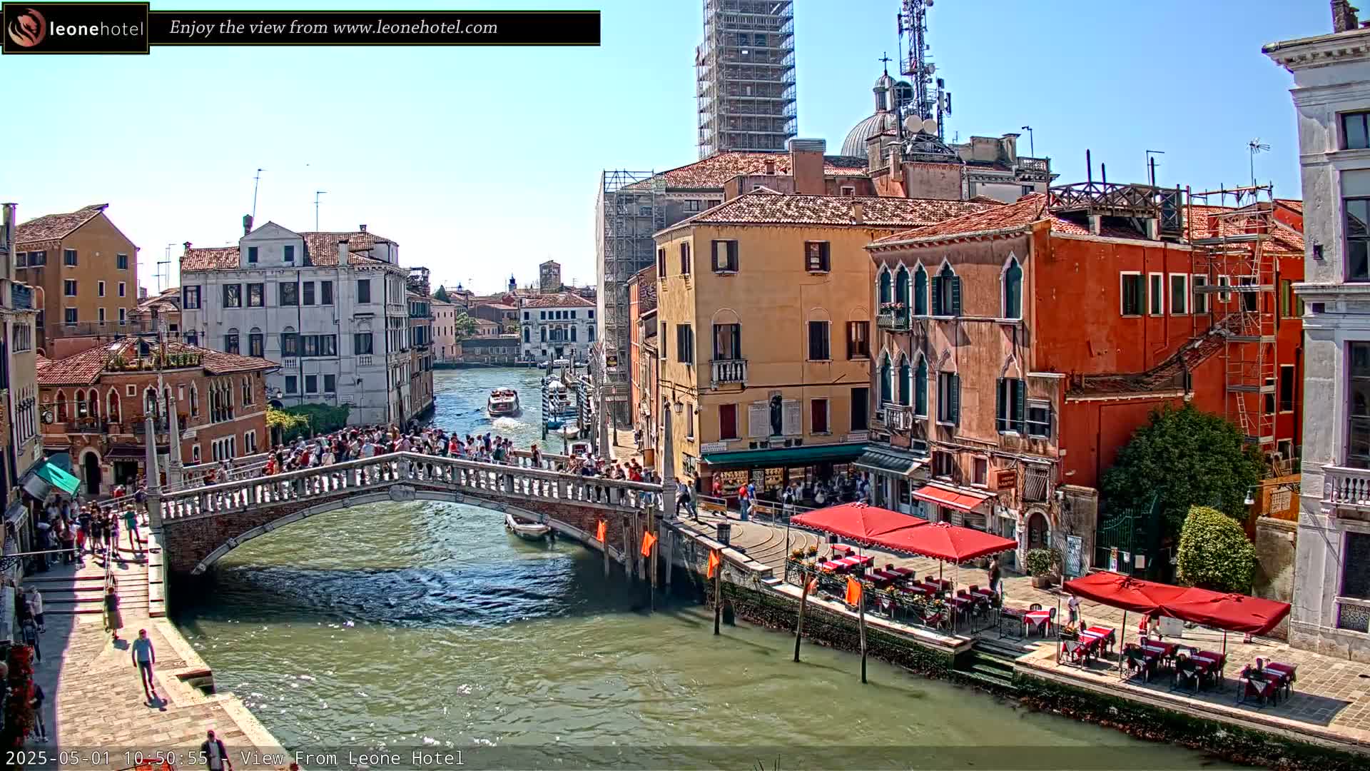 A sunny day in Venice shows a canal with boats, a bridge crowded with people, and colorful buildings lining the waterway, some with outdoor cafes.