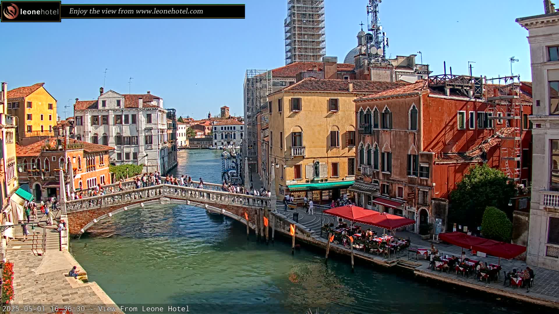 A sunny day in Venice shows a canal with a bridge crowded with people, colorful buildings lining the waterway, and outdoor cafes with red umbrellas.