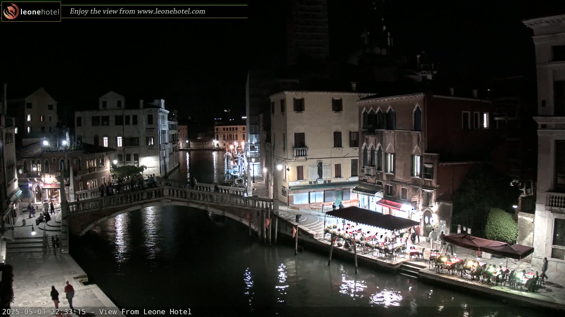 A nighttime view of a canal in Venice, Italy, showing bridges, buildings with lights, and outdoor seating at a restaurant.