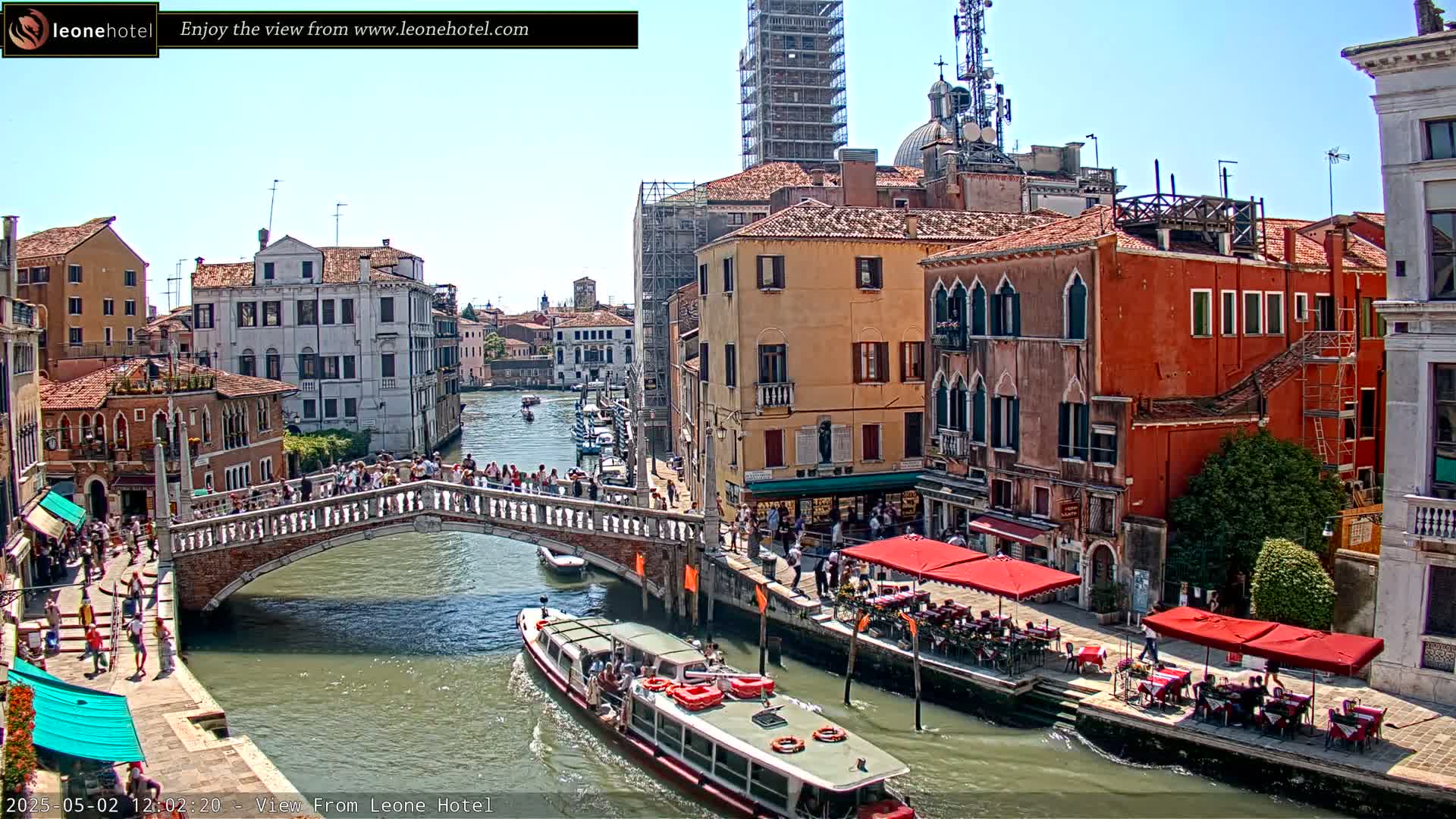 A sunny day in Venice shows a canal with a bridge crowded with people, boats, and waterfront restaurants under red umbrellas.