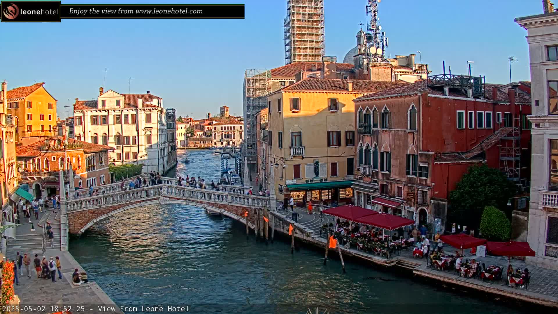 A Venetian canal scene under a clear, sunny sky shows a stone bridge crowded with pedestrians spanning a waterway lined with colorful buildings, some with outdoor cafes.