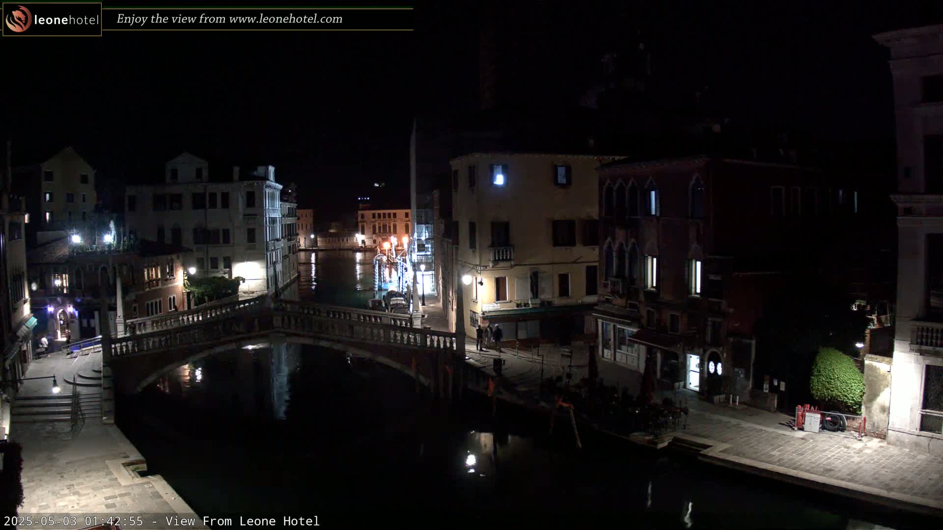 A nighttime view of a canal in Venice, Italy, shows a bridge, buildings lining the waterway, and a few people on the street.