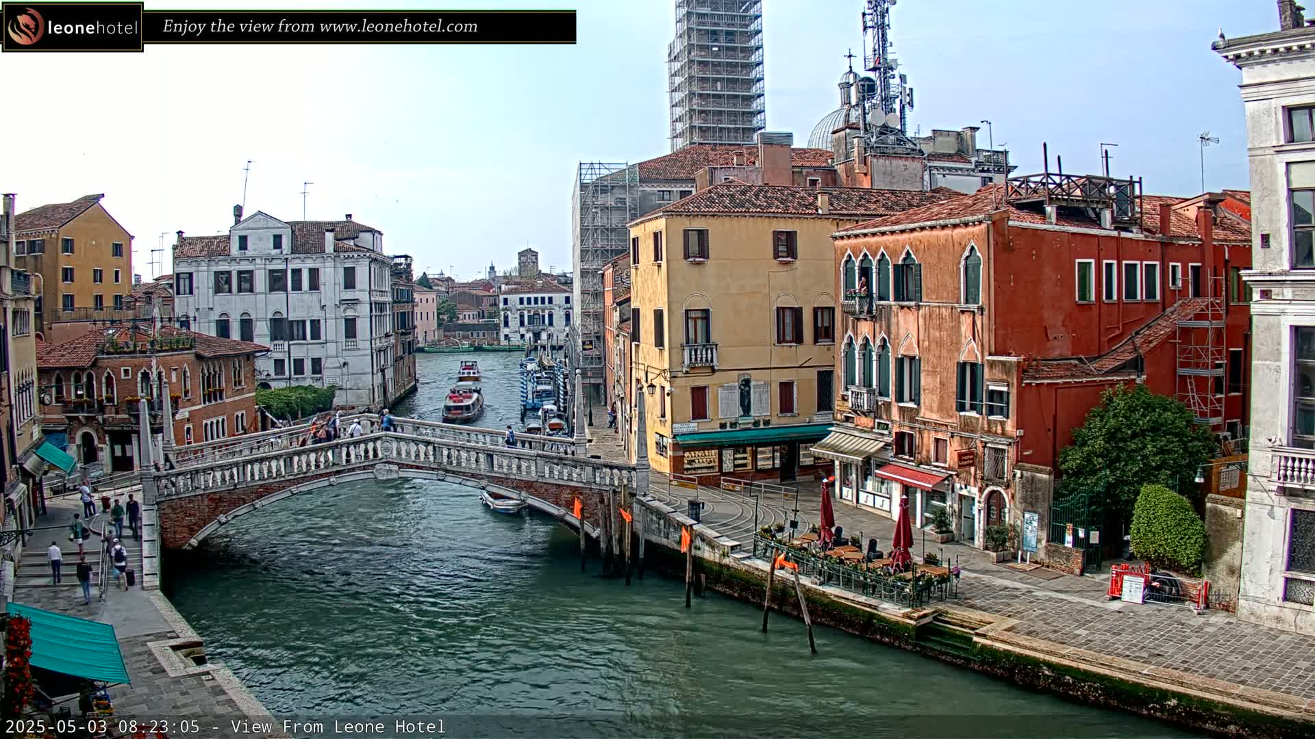 A canal in Venice, Italy, on a partly cloudy day, shows several bridges, boats, and colorful buildings lining the waterway, with people walking along the canal's edge.