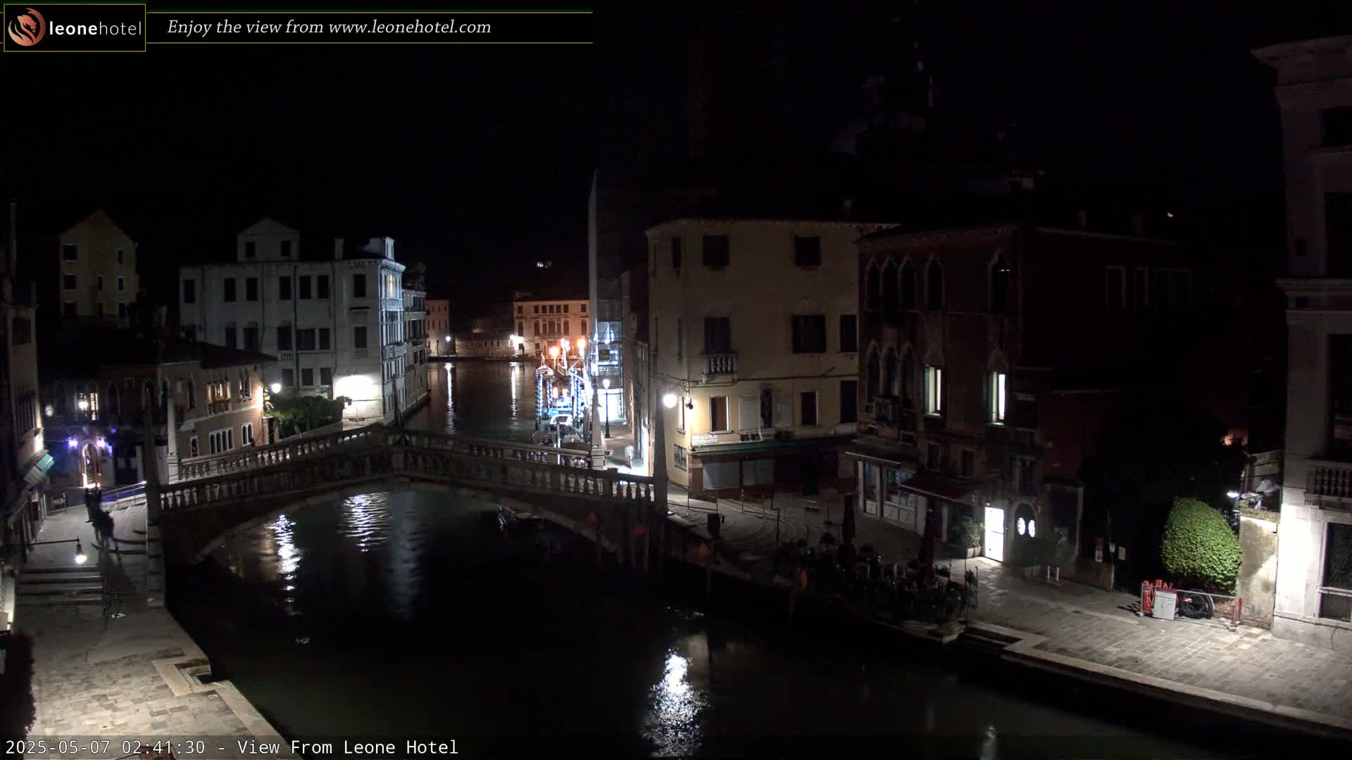 A nighttime view of a canal in Venice, Italy, shows several buildings lining the waterway, a bridge spanning the canal, and some people gathered near a restaurant.