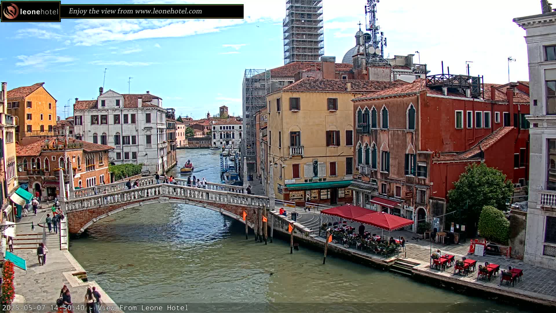 A sunny day in Venice shows a canal with a stone bridge, colorful buildings lining the waterway, and outdoor seating at a cafe.