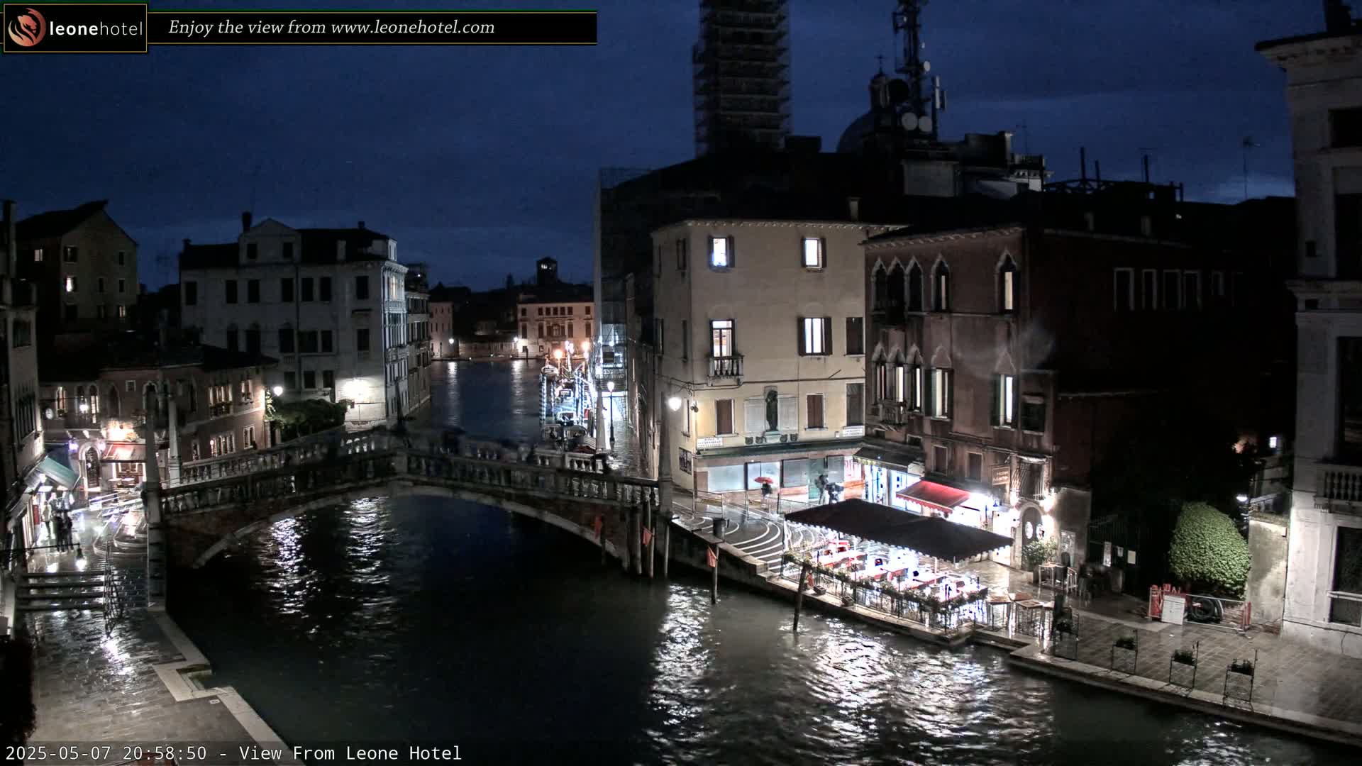 A nighttime view of a canal in Venice, Italy, showing bridges, buildings, and a restaurant with outdoor seating under a dark, possibly rainy sky.