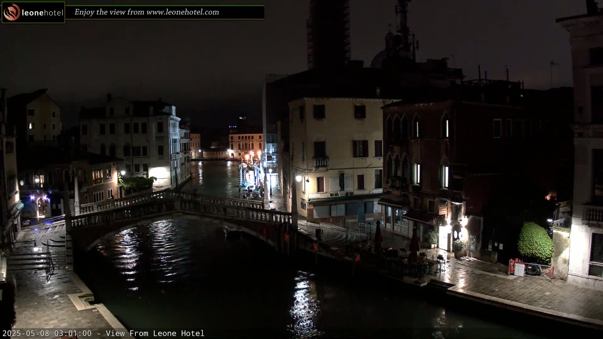 A nighttime view of a canal in Venice, Italy, shows a bridge, buildings, and wet streets under a dark sky.