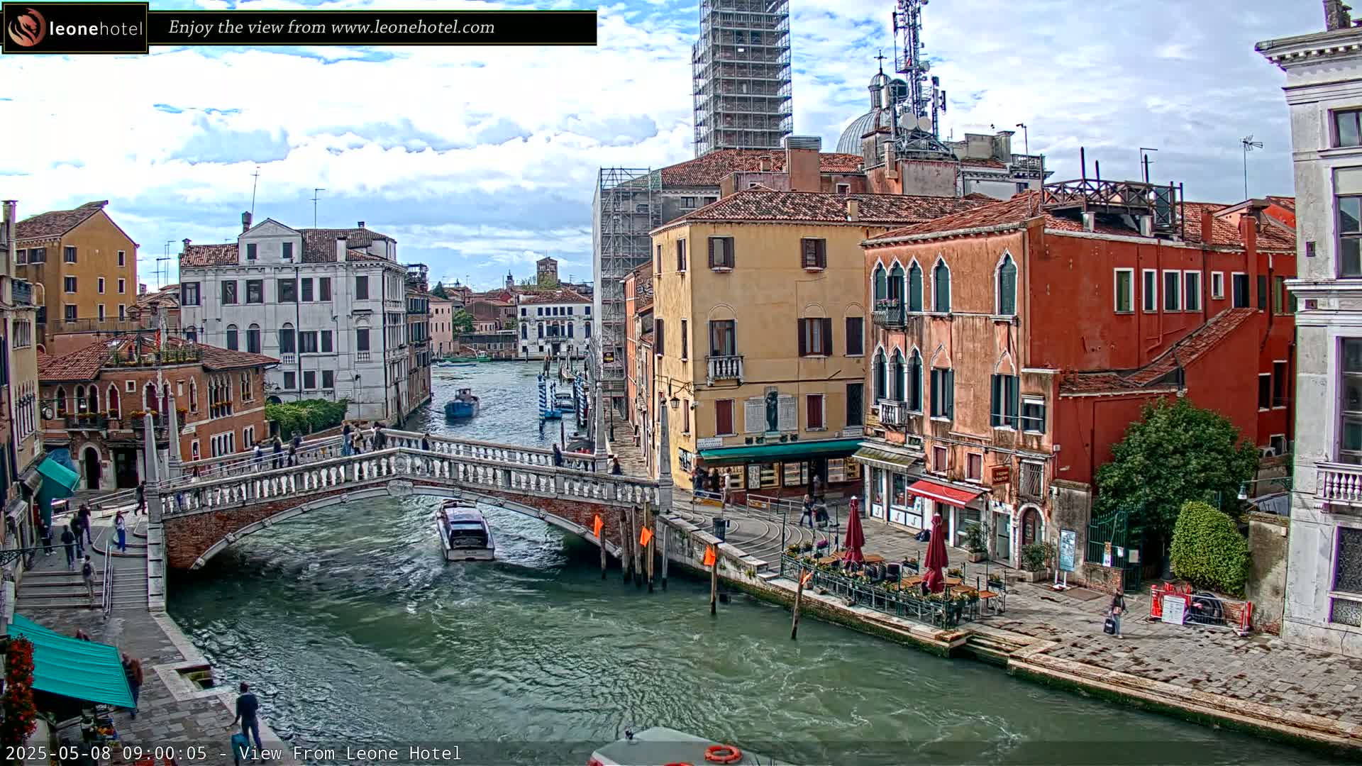 A canal in Venice, Italy, under a partly cloudy sky, shows several boats on the water, pedestrians crossing a stone bridge, and colorful buildings lining the waterway.