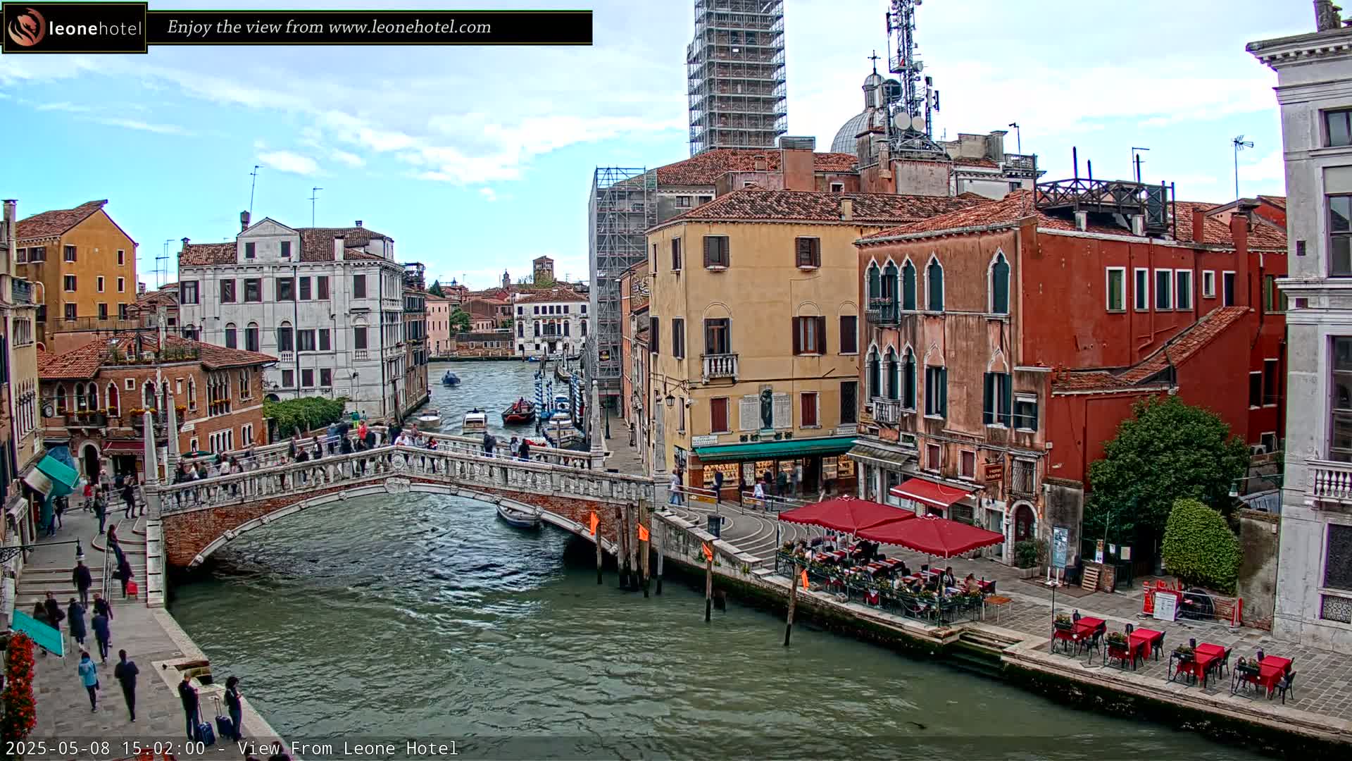 A Venetian canal scene on a partly cloudy day shows a stone bridge over the water with pedestrians crossing it, and colorful buildings lining the waterway, some with outdoor seating.