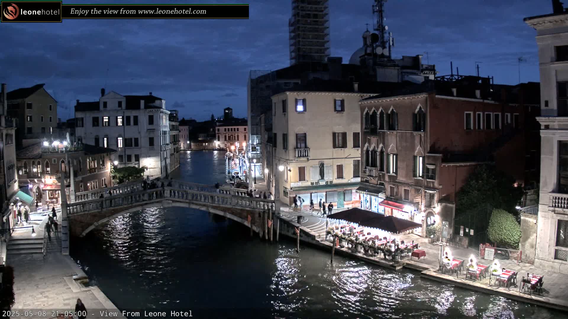 A nighttime view of a canal in Venice, Italy, shows several buildings lining the waterway, a bridge spanning the canal, and outdoor dining areas along the water's edge under a twilight sky.