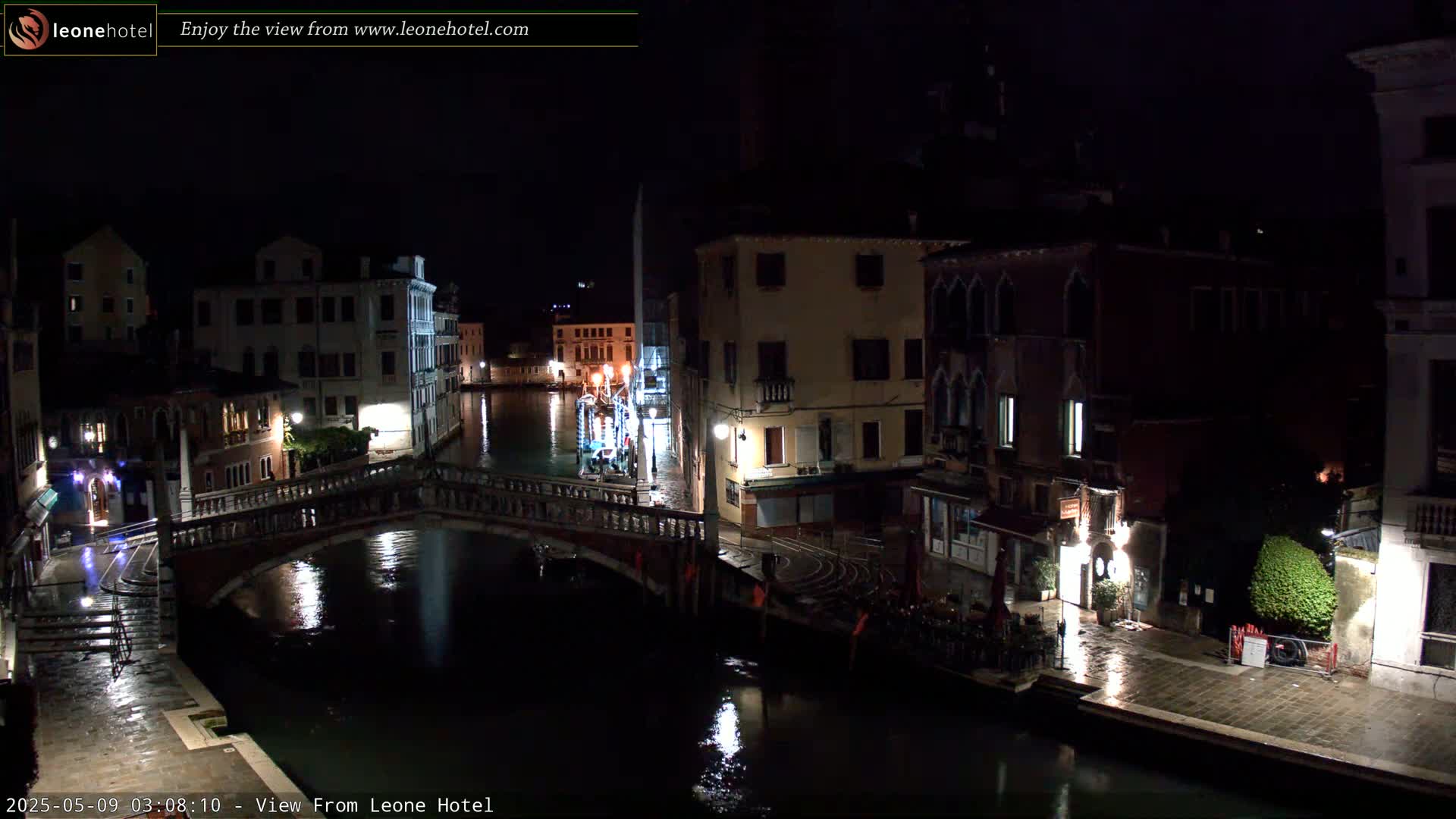 A nighttime view of a canal in Venice, with a bridge and buildings lining the waterway, under a dark, wet sky.