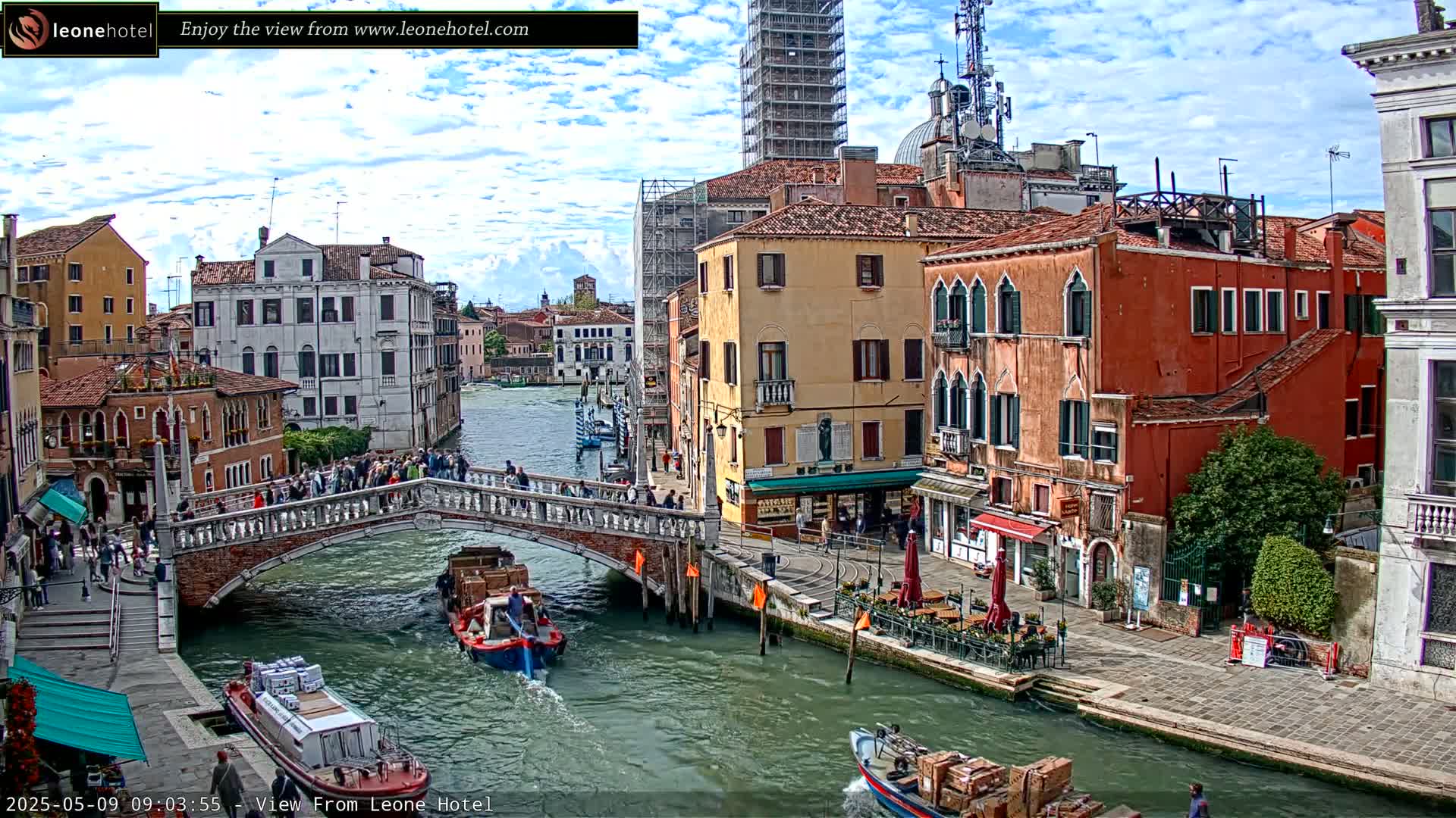 A Venetian canal scene on a partly cloudy day shows colorful buildings lining the waterway, pedestrians crossing a bridge, and boats carrying goods.