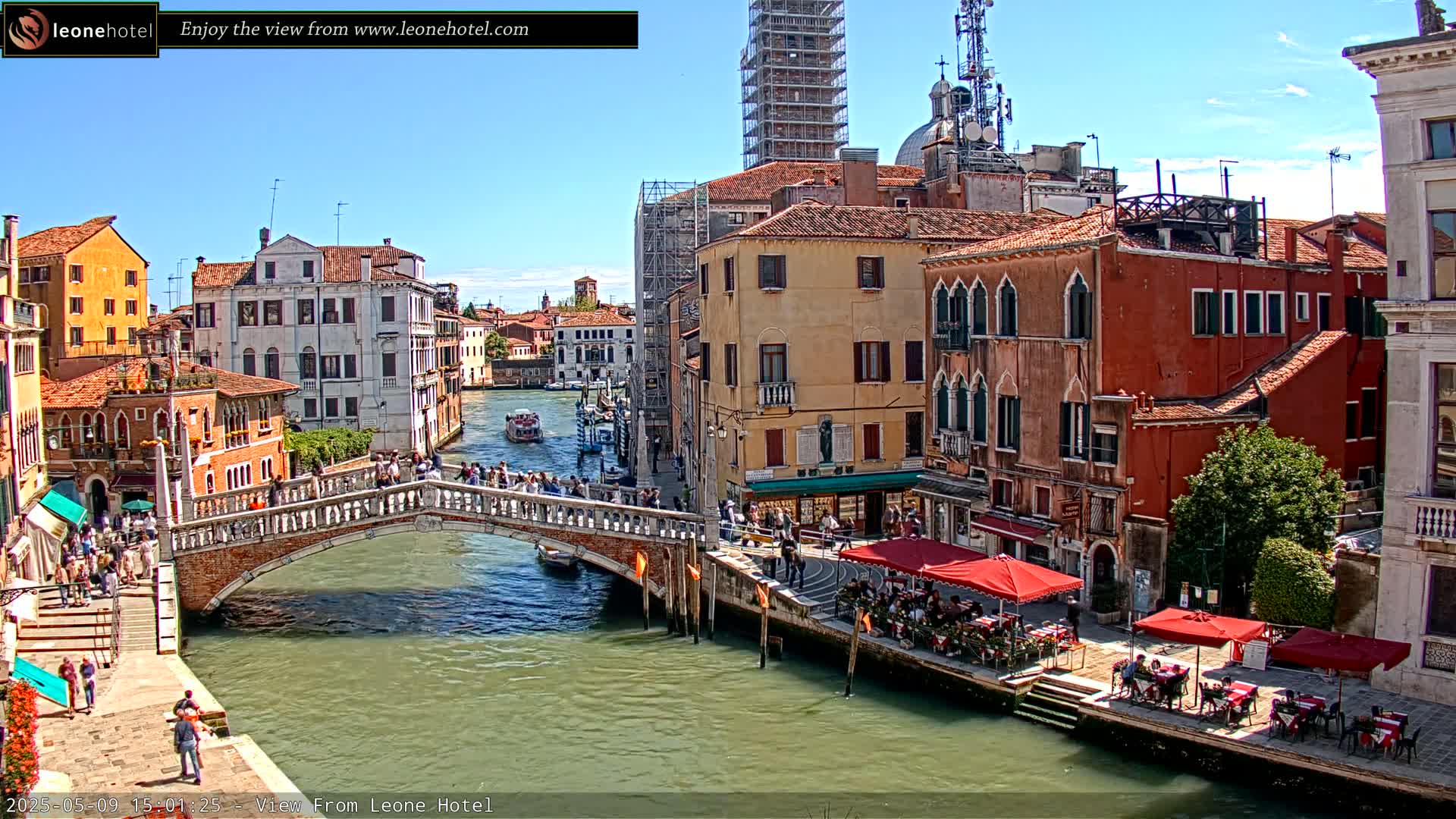 A sunny day in Venice shows a canal with a stone bridge crowded with people, boats on the water, and colorful buildings lining the waterway, with outdoor cafes along the edge.
