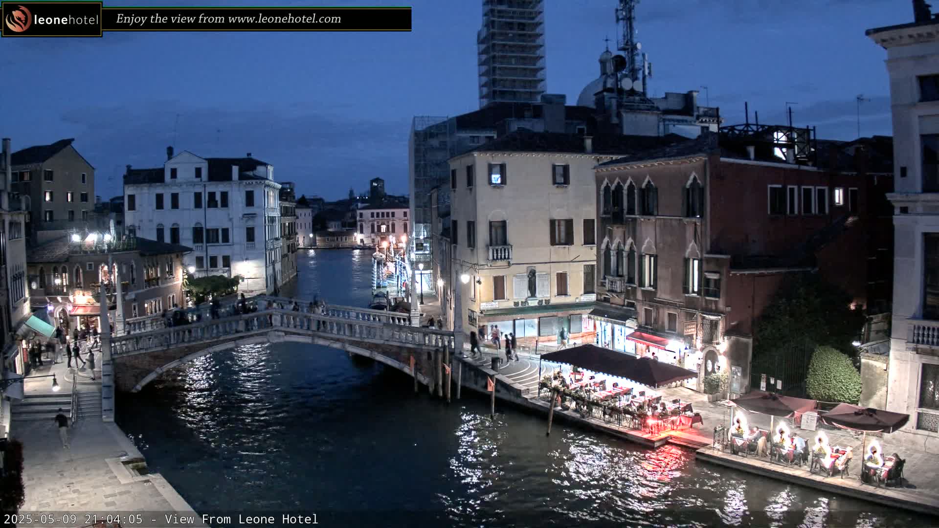 A nighttime view of a canal in Venice, Italy, showing a stone bridge, buildings with lights, and outdoor seating at restaurants along the water's edge.