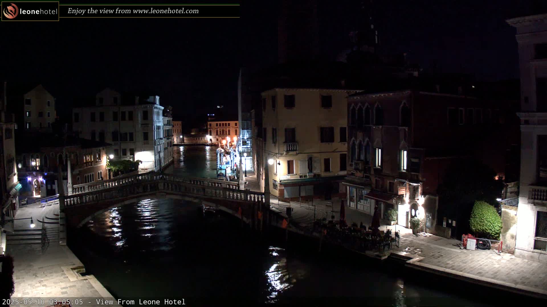 A nighttime view of a canal in Venice, with bridges, buildings, and some people gathered along the waterfront.