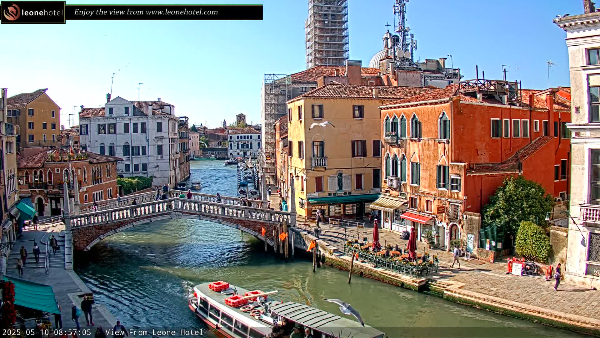 A sunny view of a canal in Venice, Italy, shows a stone bridge, colorful buildings lining the waterway, and a boat on the canal.