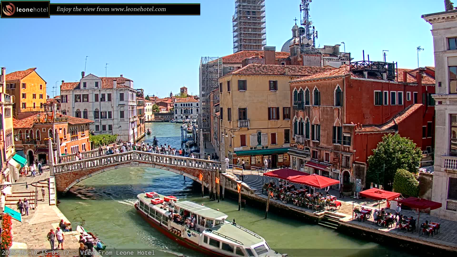 A sunny day in Venice shows a canal with a bridge full of people, boats, and buildings lining the waterway, including outdoor cafes.