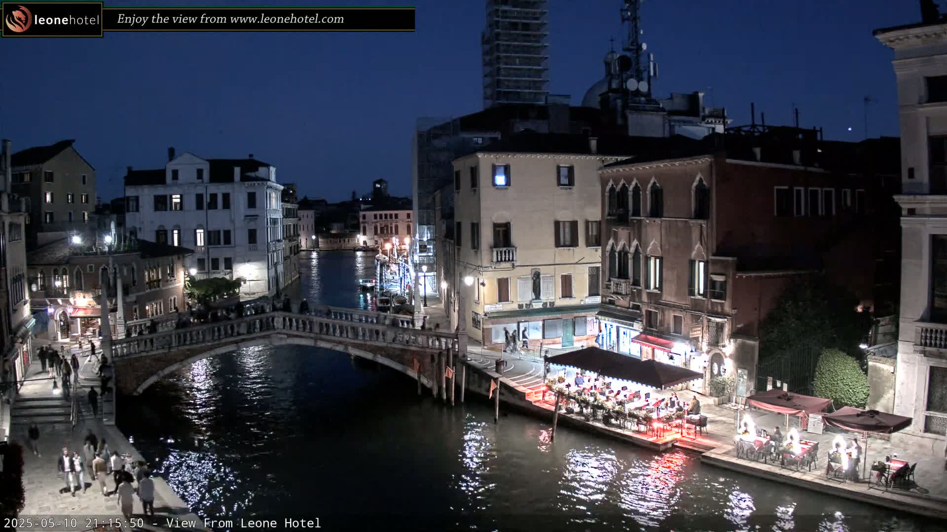 A nighttime view of a canal in Venice, Italy, shows a bridge with pedestrians, buildings with lights, and outdoor dining areas along the water's edge.