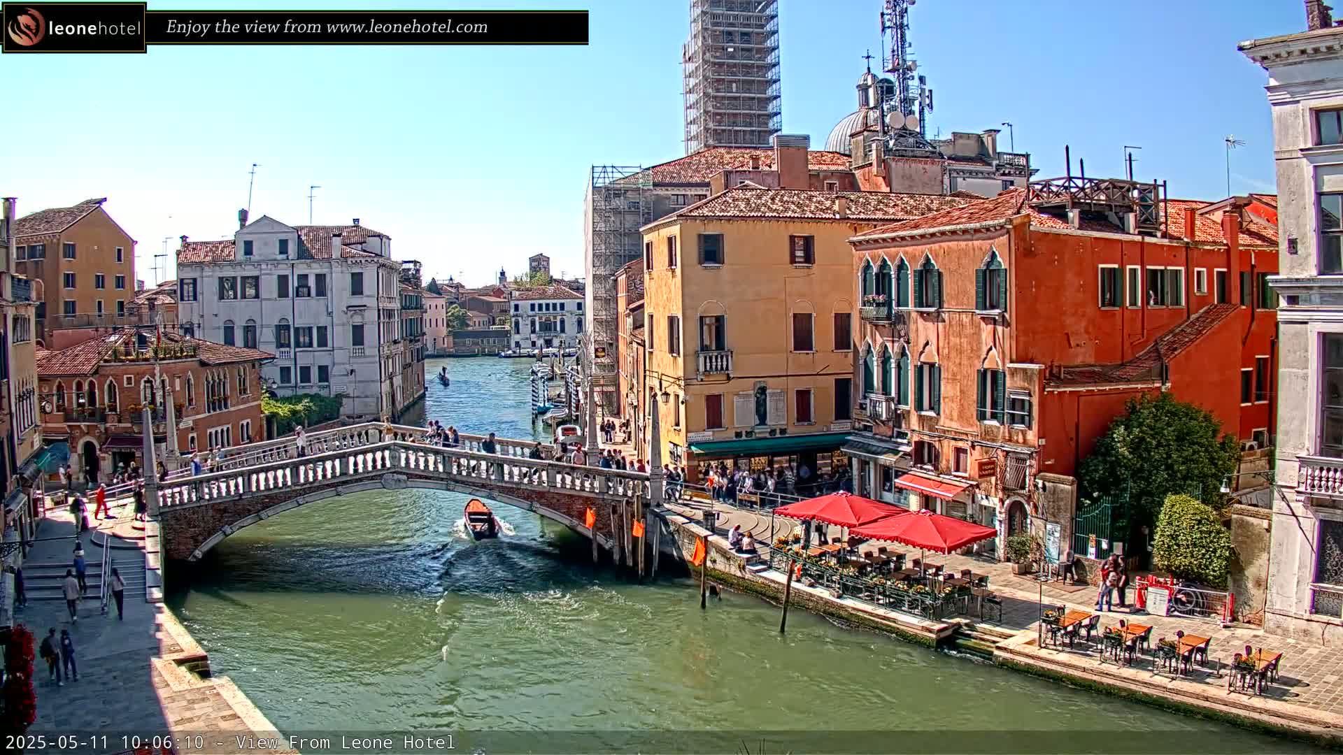 A sunny day in Venice shows a canal with a bridge, boats, and colorful buildings lining the waterway, with outdoor seating at a cafe.