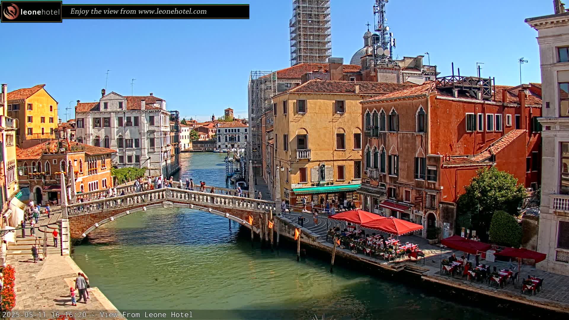 A sunny day in Venice shows a canal with a stone bridge, colorful buildings lining the waterway, and outdoor seating at restaurants.