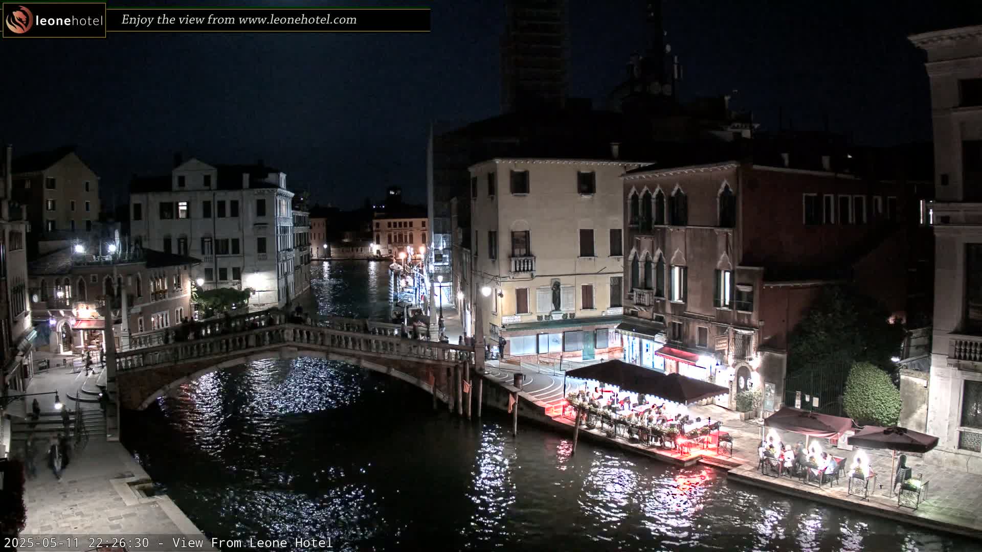 A nighttime view of a canal in Venice, Italy, shows several buildings lining the waterway, a bridge crossing the canal, and outdoor dining areas with people seated at tables.