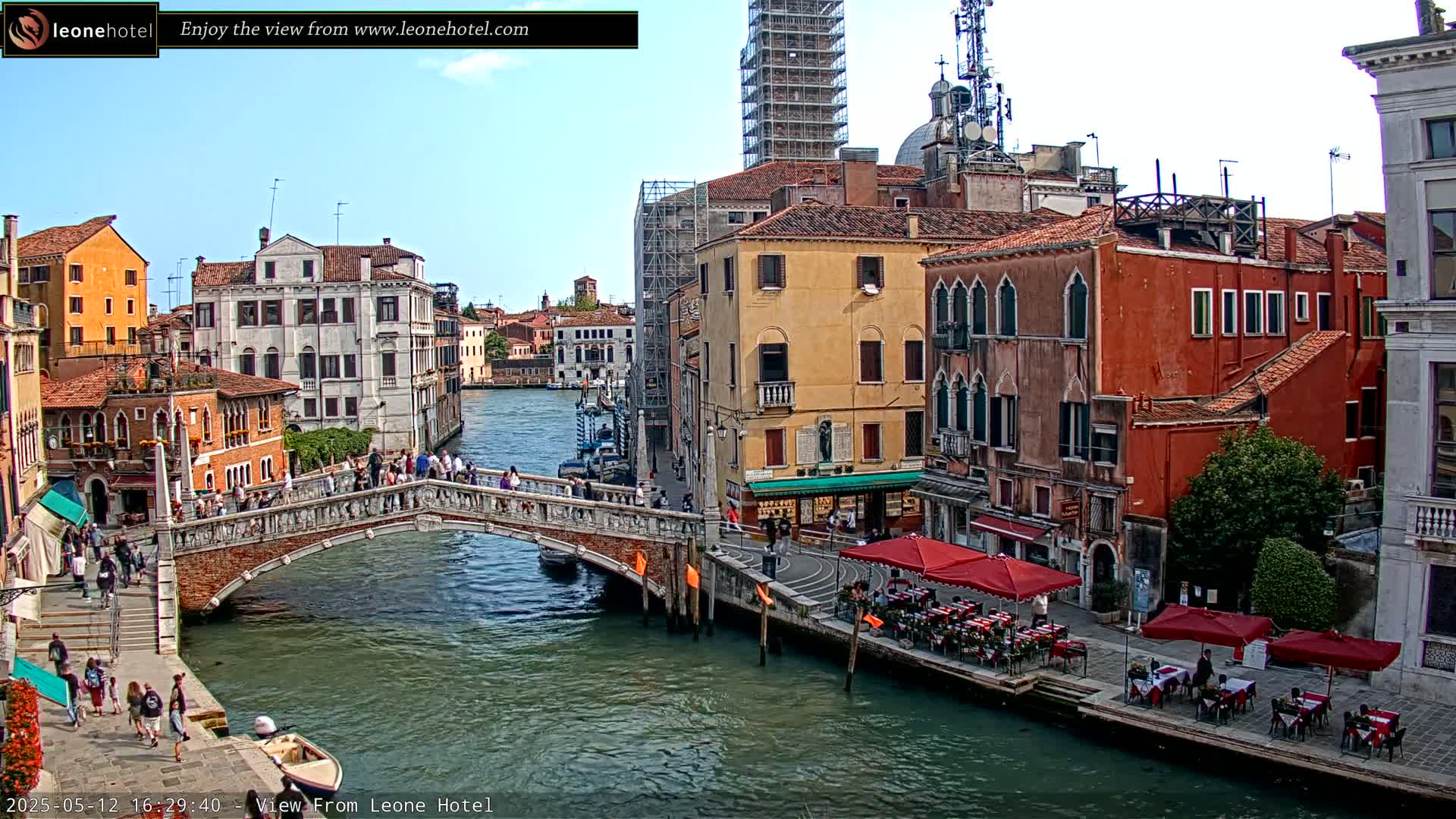 A sunny day in Venice shows a canal with a bridge, several buildings along the waterway, and outdoor seating at cafes.