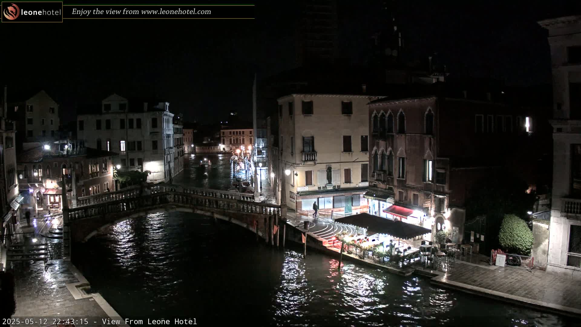 A nighttime view of a canal in Venice, Italy, shows bridges, buildings, and a restaurant with outdoor seating, under a dark sky.