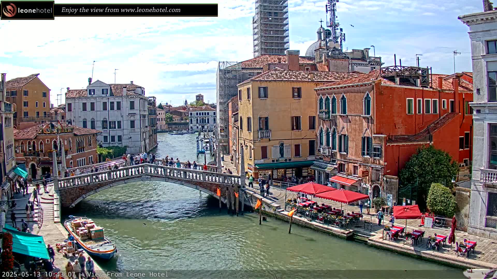 A stone bridge over a canal in Venice, Italy, on a partly cloudy day, is lined with people and has buildings with orange and tan facades along its sides.