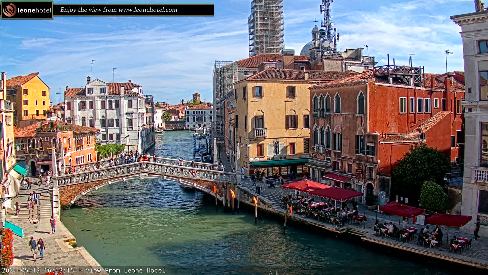 A sunny day in Venice shows a canal with a stone bridge, colorful buildings lining the waterway, and outdoor seating at cafes along the banks.