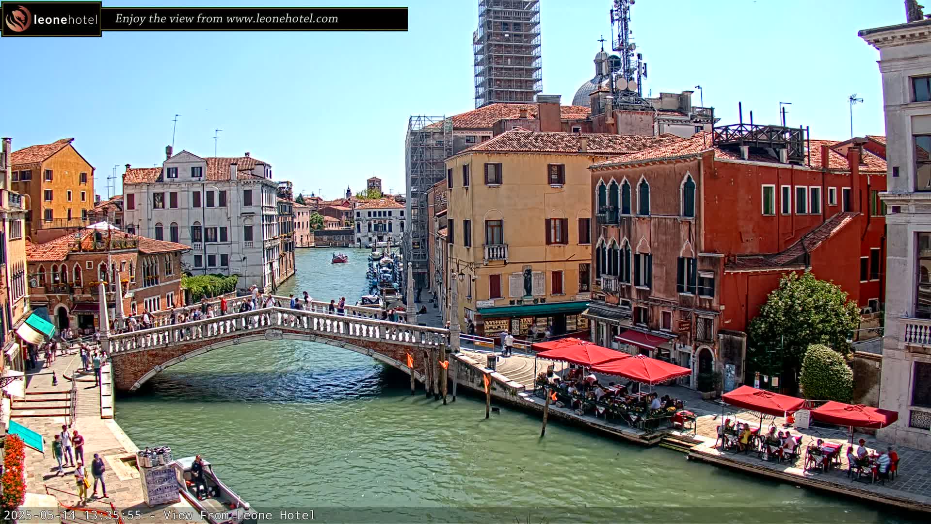 A sunny day in Venice shows a canal with a stone bridge, colorful buildings lining the waterway, and outdoor seating at cafes.